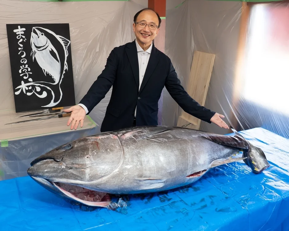 笑顔の男性が巨大なマグロを指し示している。背景には「まぐろの学」と書かれた看板があり、マグロの解体準備の様子を捉えた写真である。