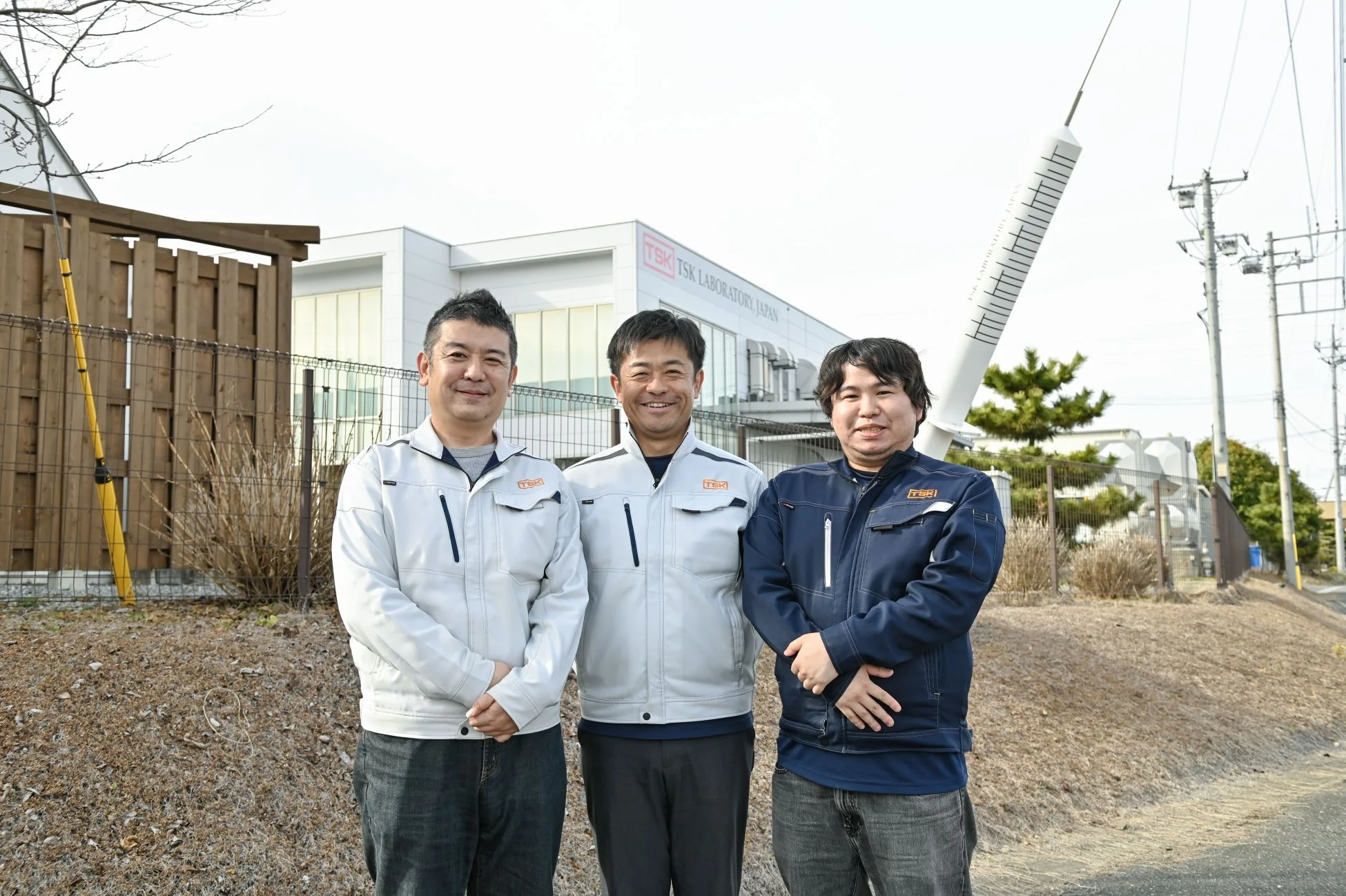 TSK LABORATORY JAPANの建物前で作業着姿の男性3人が笑顔で立っています。背景には巨大な注射器のモニュメントも見え、研究施設であることが伺えます。