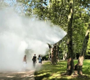 公園の木々の間を、人々が白い霧の中を歩いている様子