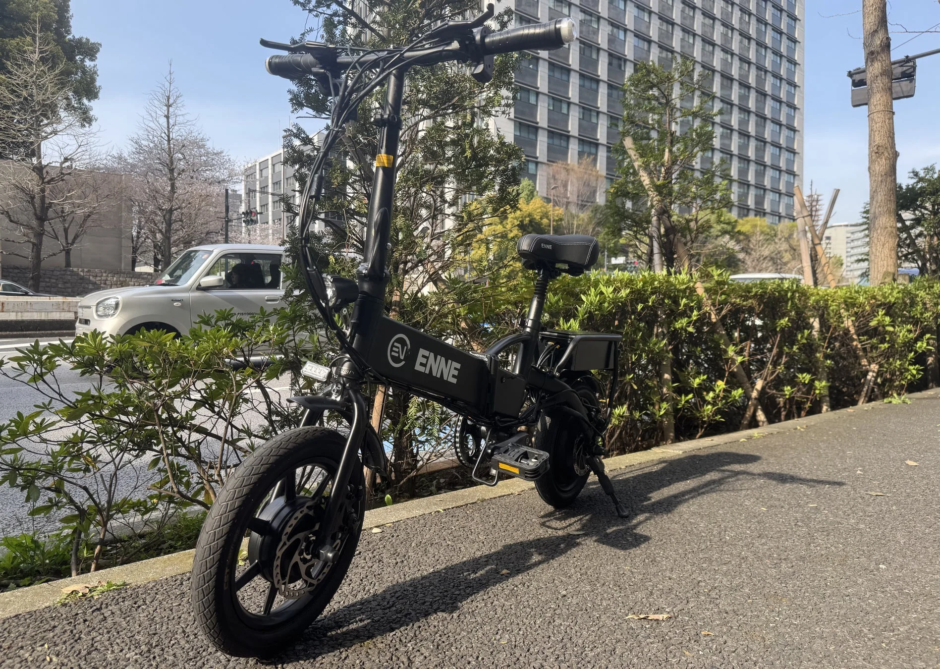 晴れた日の歩道に停められた黒い電動折りたたみ自転車「ENNE」が写っています。背景にはビルや車、木々が見え、都市の日常風景が広がっています。