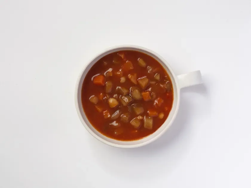 白い背景に置かれた白いボウルに入った、にんじんなどの野菜がたっぷり入った具沢山のトマトベースのスープまたはシチューです。温かそうな一品が上から撮影されています。