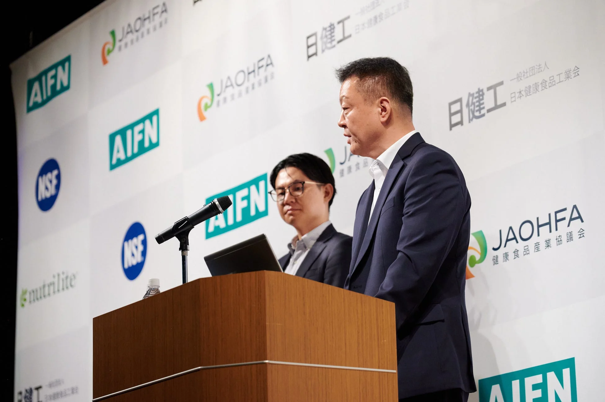 健康食品関連のロゴが背景に並ぶ会場で、スーツ姿の男性がマイクを使ってプレゼンテーションを行っている様子