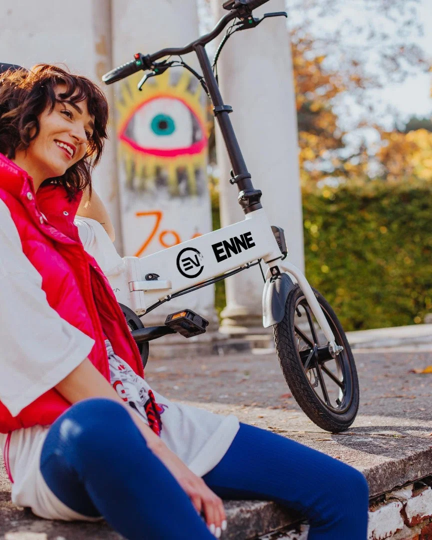 ピンクのベストと青いパンツを着用した若い女性が、白い折りたたみ式電動自転車の隣に座って笑顔を見せています。背景にはグラフィティのある壁と秋の木々が見え、リラックスした屋外の雰囲気が伝わります。