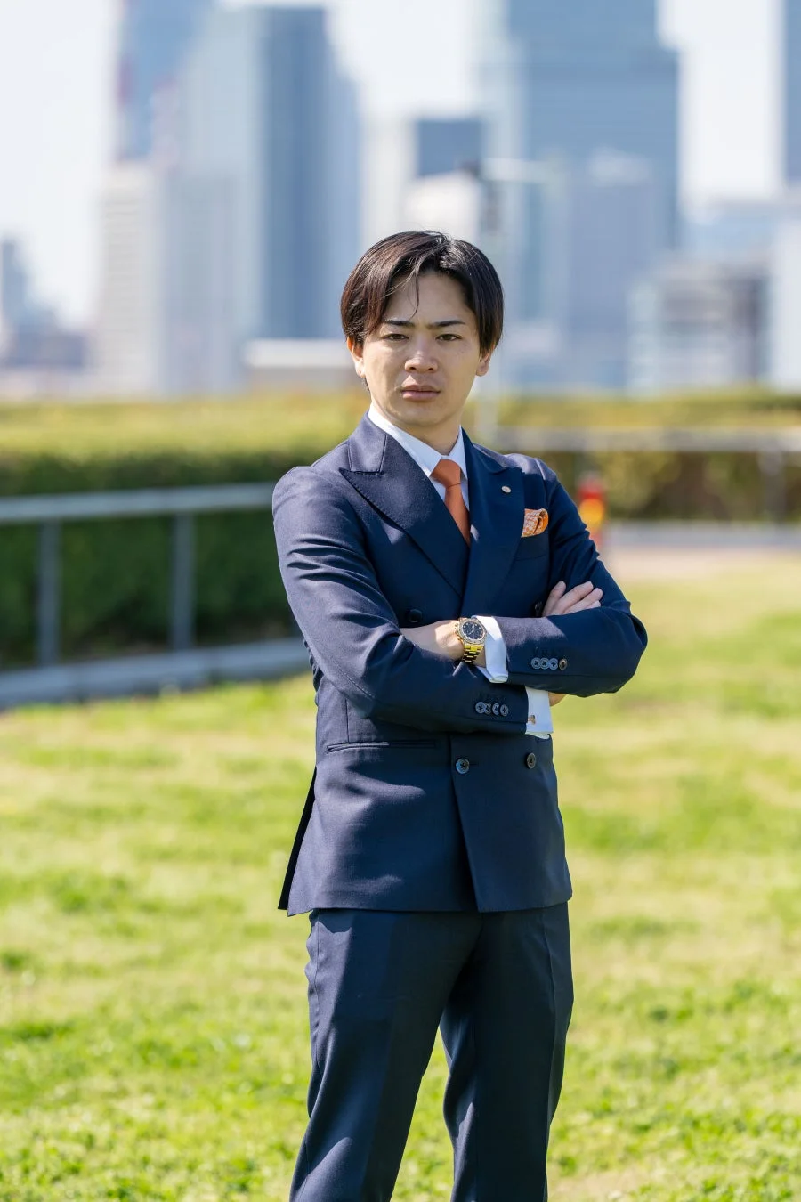 紺色のダブルスーツを着た若い日本人男性が、腕を組んで自信に満ちた表情で立っています。背景には芝生とぼやけた都市の建物が広がり、ビジネスシーンを思わせるポートレートです。