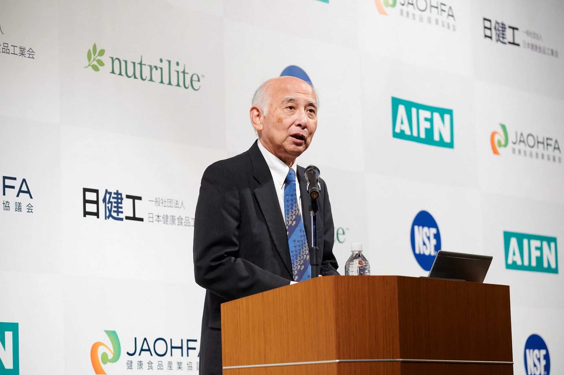 スーツを着た高齢の男性が、企業ロゴが並ぶ背景の前で演台に立ち、スピーチをしている様子