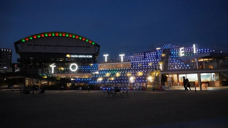 夜の神戸のウォーターフロントにあるGLION ARENA KOBEと、鮮やかにライトアップされた「TOTTEI」の文字が見える施設を捉えた画像です。階段状の構造物には無数の電飾が施され、幻想的な雰囲気を醸し出しています。