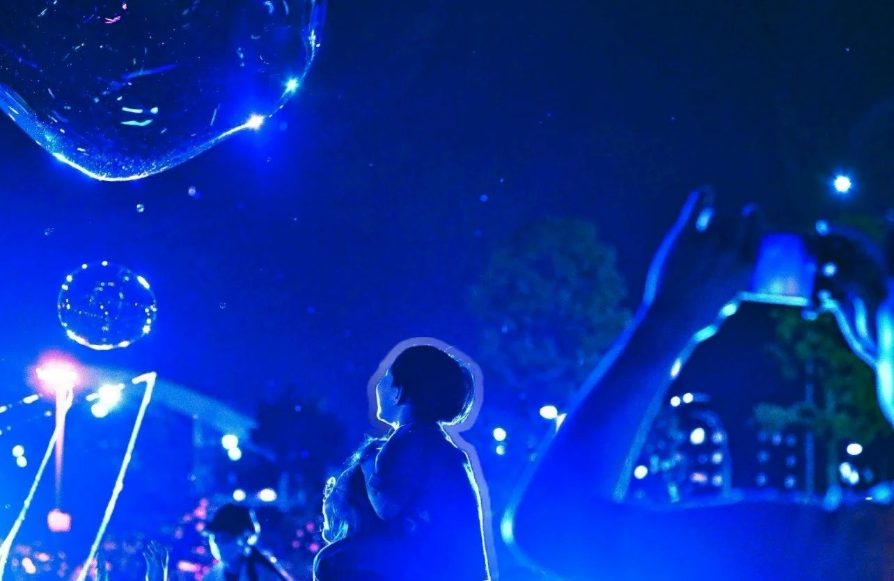 青い光に包まれた夜のイベントで、人々が空に舞うシャボン玉を見上げている様子