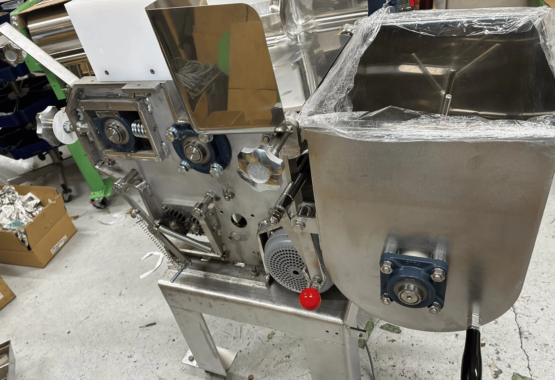 工場のような場所に置かれたステンレス製の工業機械です。ローラーやギア、モーター、ホッパーなどの部品が見られ、食品加工機と推測されます。一部ビニールで覆われており、新品または未使用の状態を示唆しています。