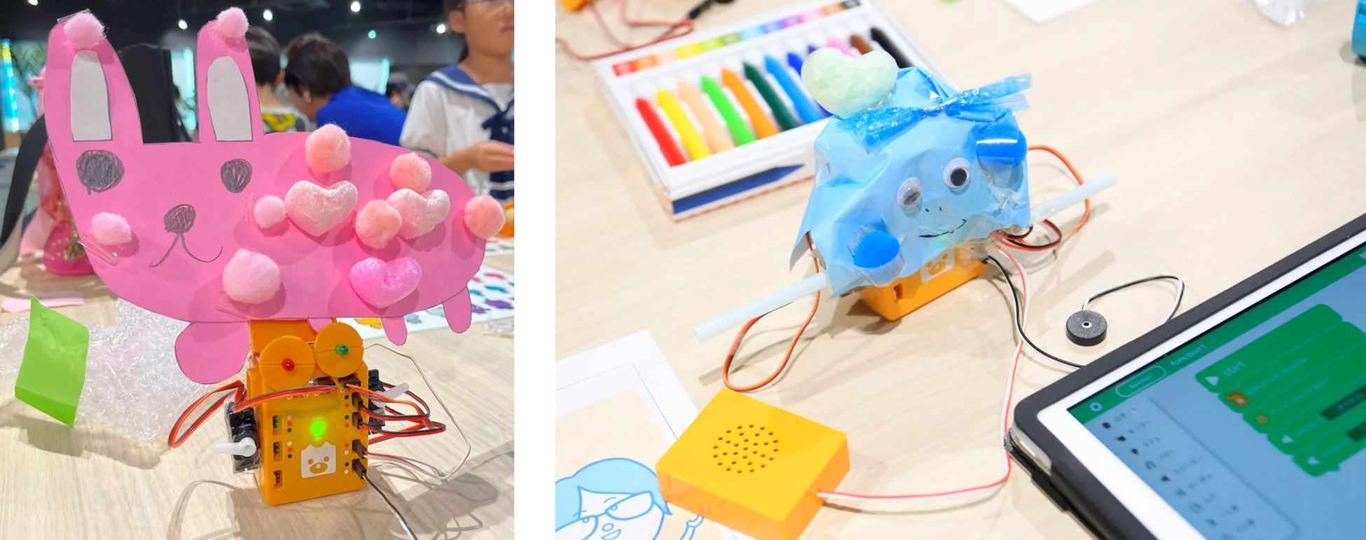 子供たちが電子部品を使ったクラフト作品を制作している様子。ピンクのウサギと青い生き物の工作が電子モジュールに接続され、プログラミング画面が表示されたタブレットや画材も見られる。