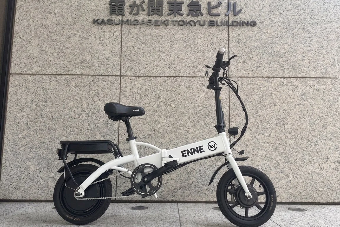 白い折りたたみ電動自転車が、霞が関東急ビルの壁を背景に停まっている様子。都市部の風景と最新の移動手段を組み合わせた一枚です。