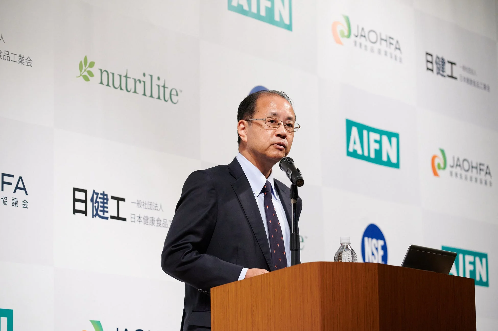 スーツを着た男性がマイクを使って壇上でスピーチをしています。背景にはnutrilite、AIFN、JAOHFA、日建工などの企業ロゴが見られ、健康食品関連のイベントの様子です。