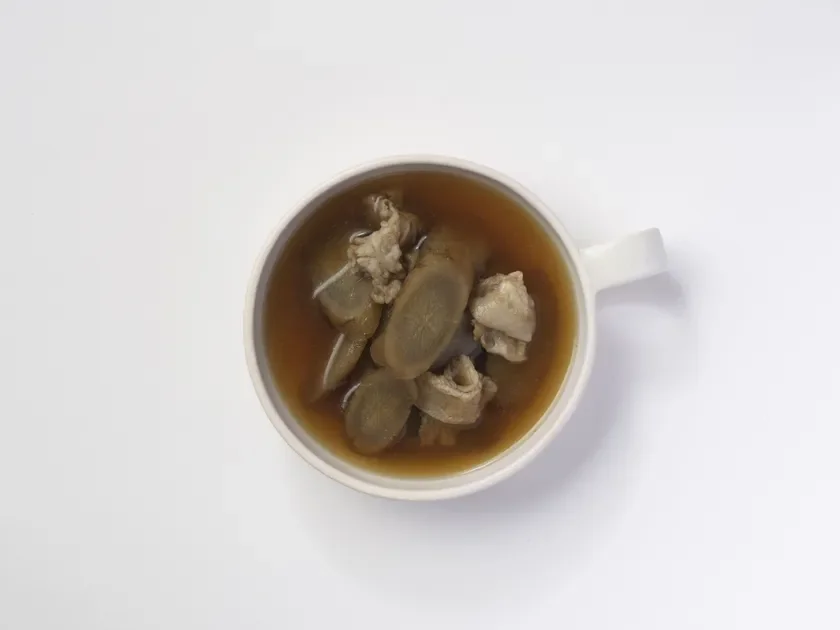 白いマグカップに、肉とスライスされた根菜が入った茶色いスープが注がれています。白い背景に置かれ、上から撮影された料理の写真です。