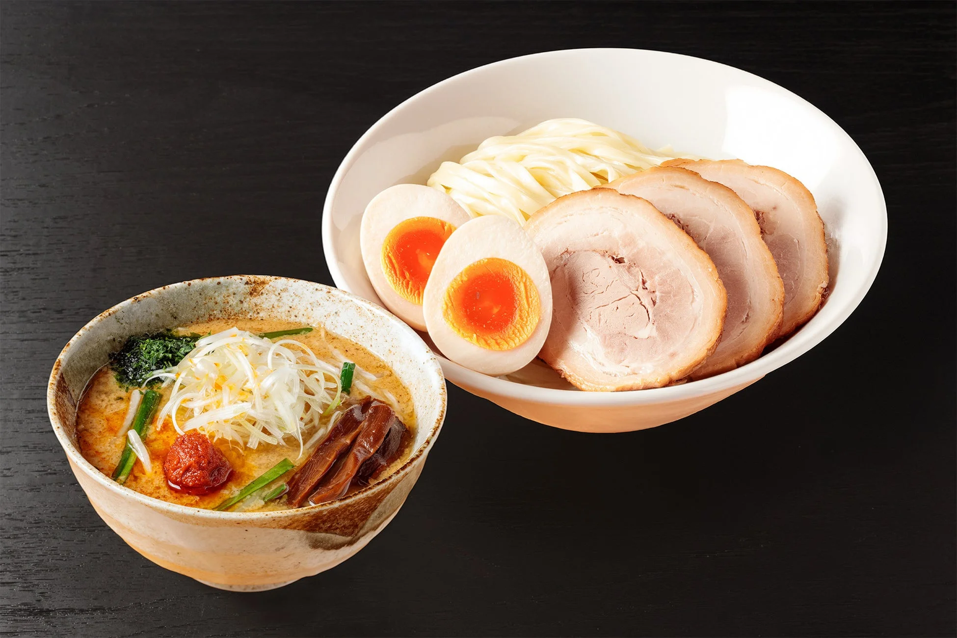 濃厚なつけ汁と太麺が特徴のつけ麺。チャーシュー3枚と半熟卵2個が添えられた麺、そしてネギ、メンマ、辛味噌が彩り豊かなつけ汁の器が並んでいます。食欲をそそる一品です。