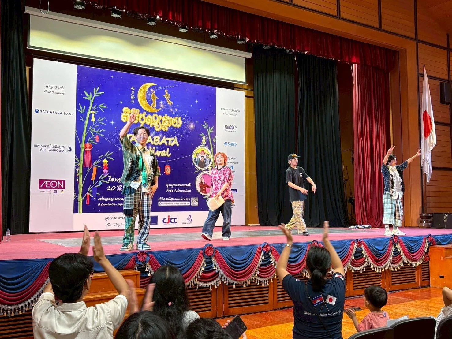 カンボジアで開催された七夕祭りのイベント風景。ステージ上の出演者が観客に手を振り、後方スクリーンにはイベント名やスポンサーロゴが表示されている。