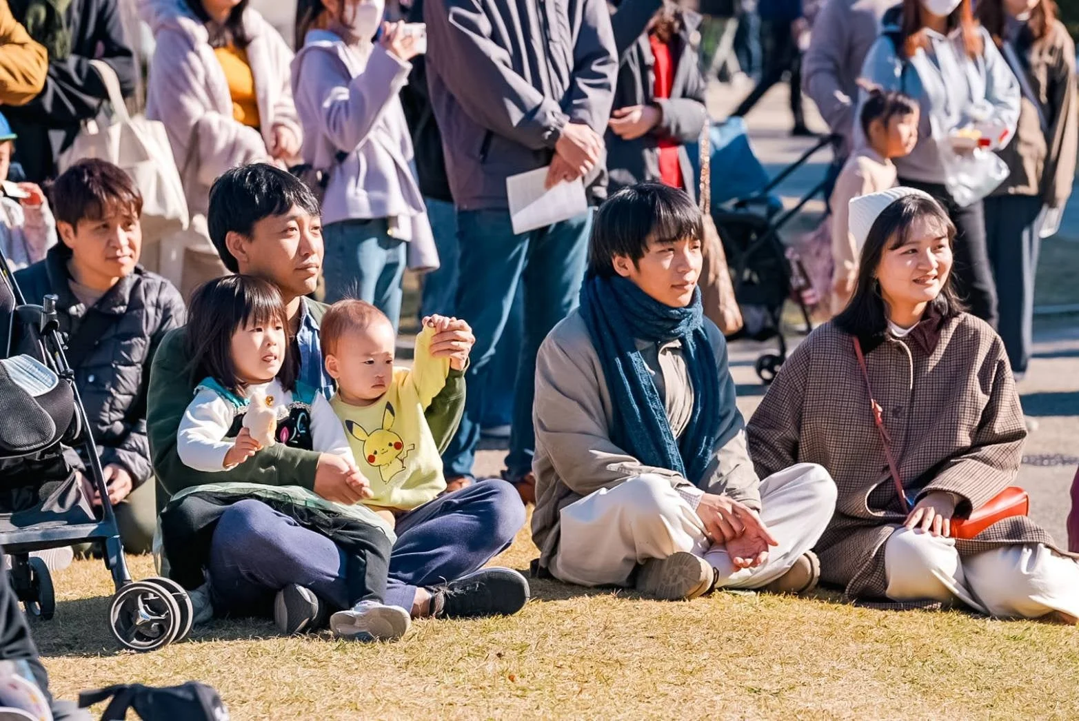 屋外の公園で、家族連れや友人たちが芝生に座ってくつろいでいる様子