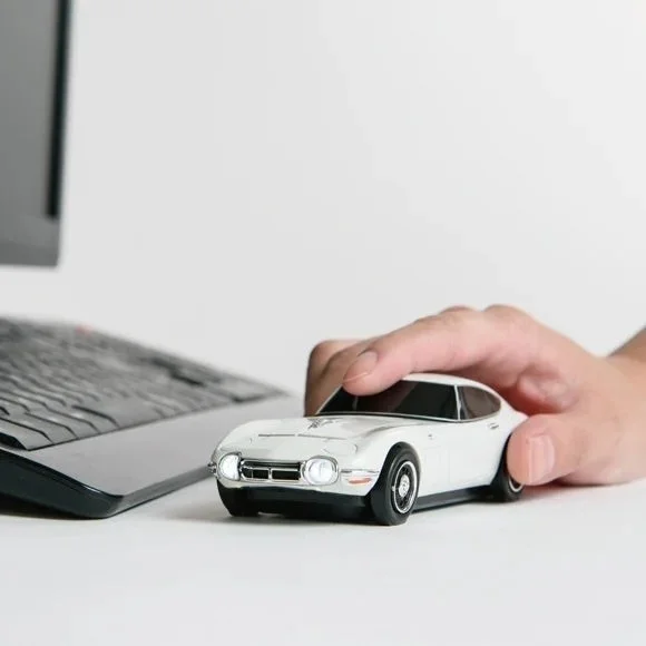 白いデスクの上で、手が白いトヨタ2000GTのミニカーを握っている様子。背景にはノートパソコンのキーボードが一部映り込んでおり、クラシックカーの模型と現代のデスク環境が融合した一枚。