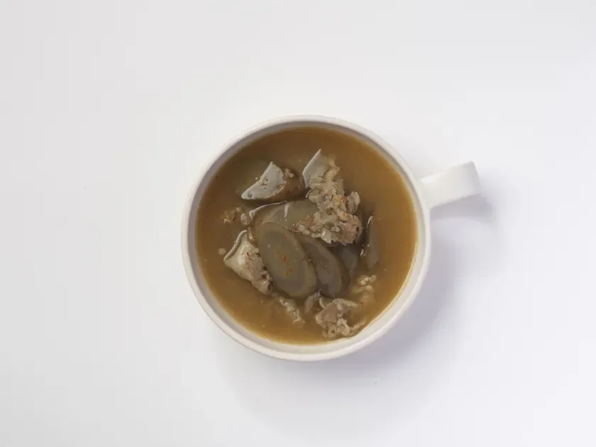 白い背景に置かれた白いマグカップに、肉とスライスされた野菜が入った茶色のスープまたは煮込み料理が盛られています。温かい食事が連想されます。