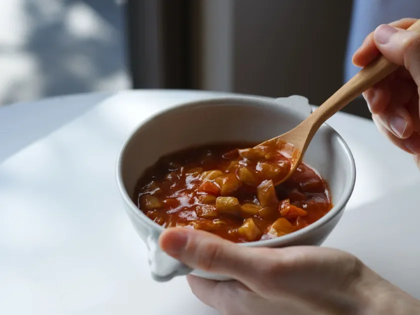 白いボウルに入った、具材が豊富な温かいシチューを、木のスプーンで食べる瞬間のクローズアップ。食欲をそそる一場面です。