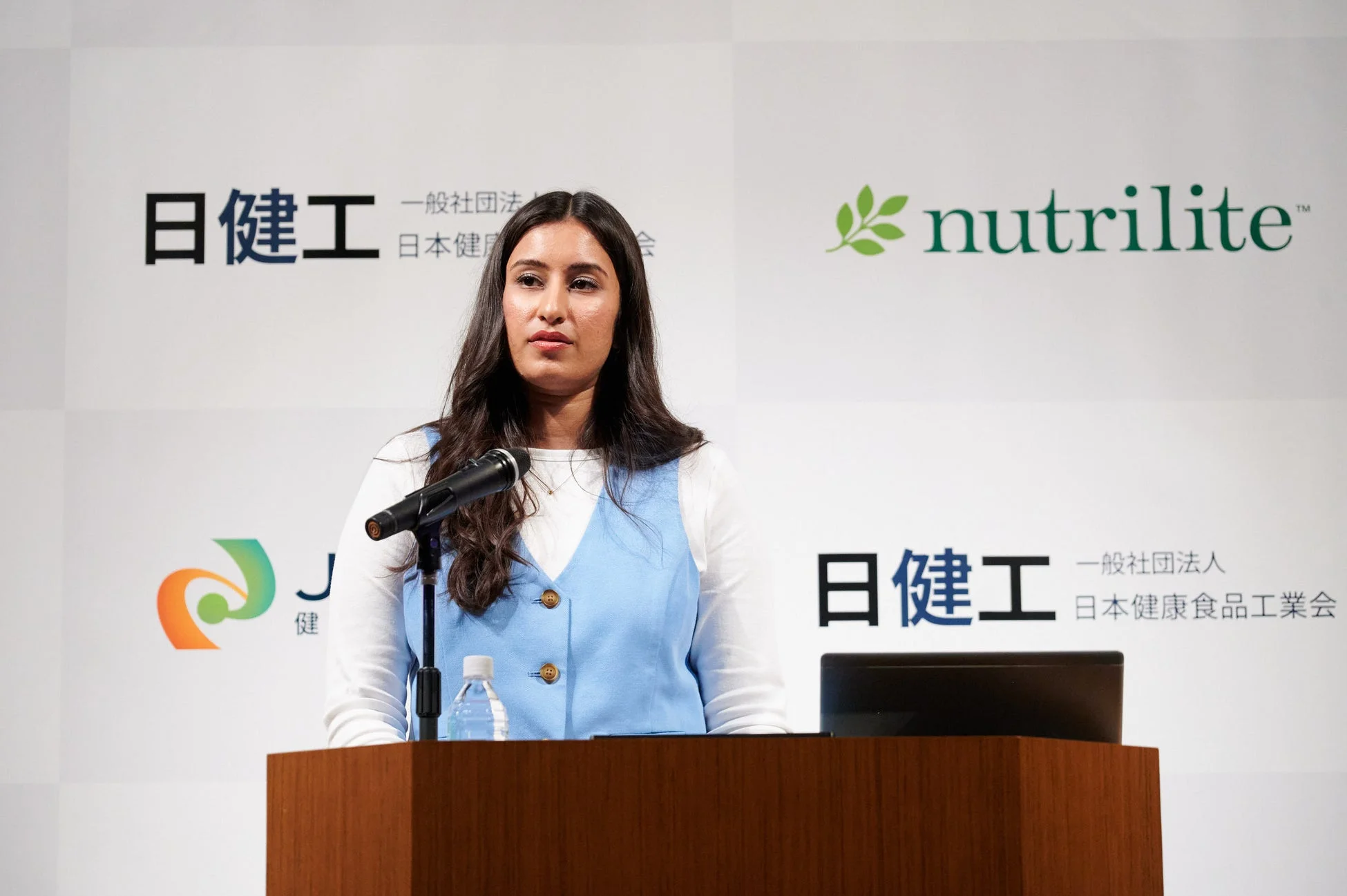 女性がマイクの前に立ち、講演を行っている様子。背景には「日建工」「一般社団法人 日本健康食品工業会」「nutrilite」のロゴと文字が見え、健康食品業界に関連するビジネスイベントと推測されます。
