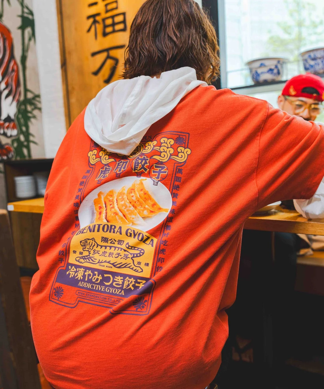 餃子のグラフィックがプリントされたオレンジ色のフーディーを着た人物の後ろ姿