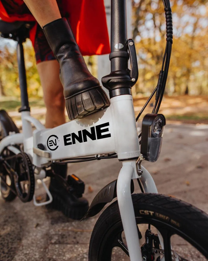白い折りたたみ電動自転車のペダルに黒いブーツを履いた人物の足が乗っているクローズアップ画像です。背景には秋の木々が見えます。