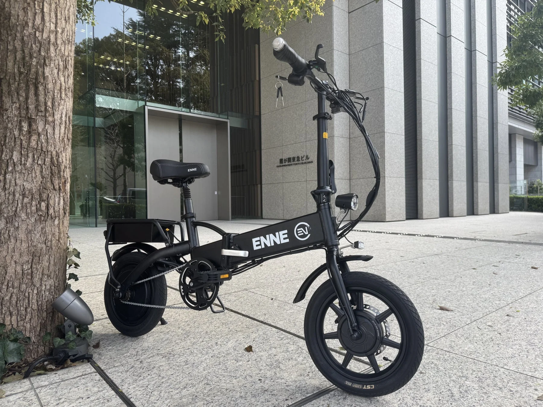 近代的なオフィスビルの前、木陰に黒いENNE EVの折りたたみ電動自転車が停まっている。都市での移動に適したスタイリッシュなデザインが特徴で、霞が関東急ビルが背景に見える。