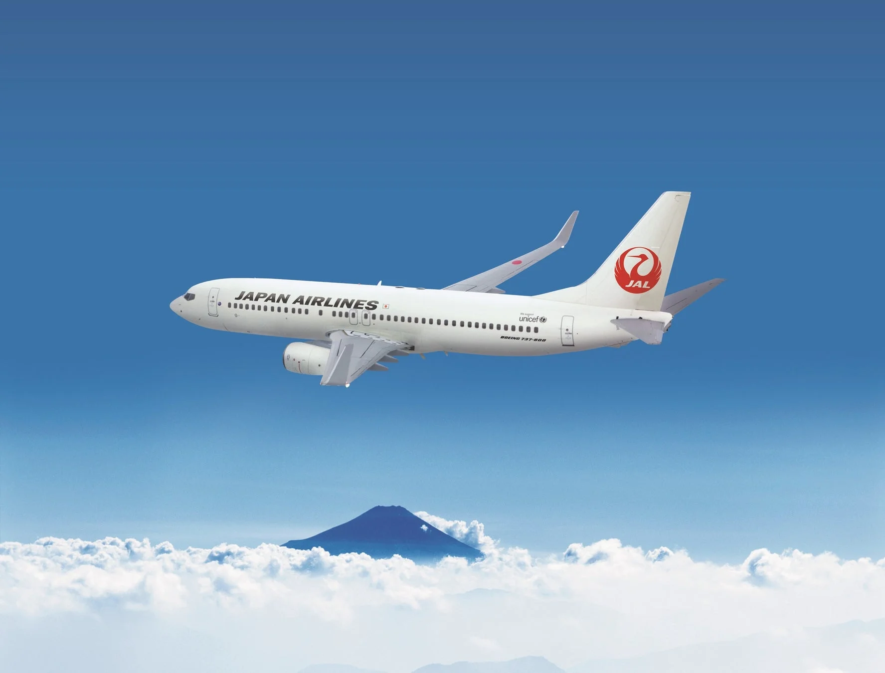 JALの飛行機が青空と雲の上を飛行しており、背景には富士山が見える雄大な風景です。
