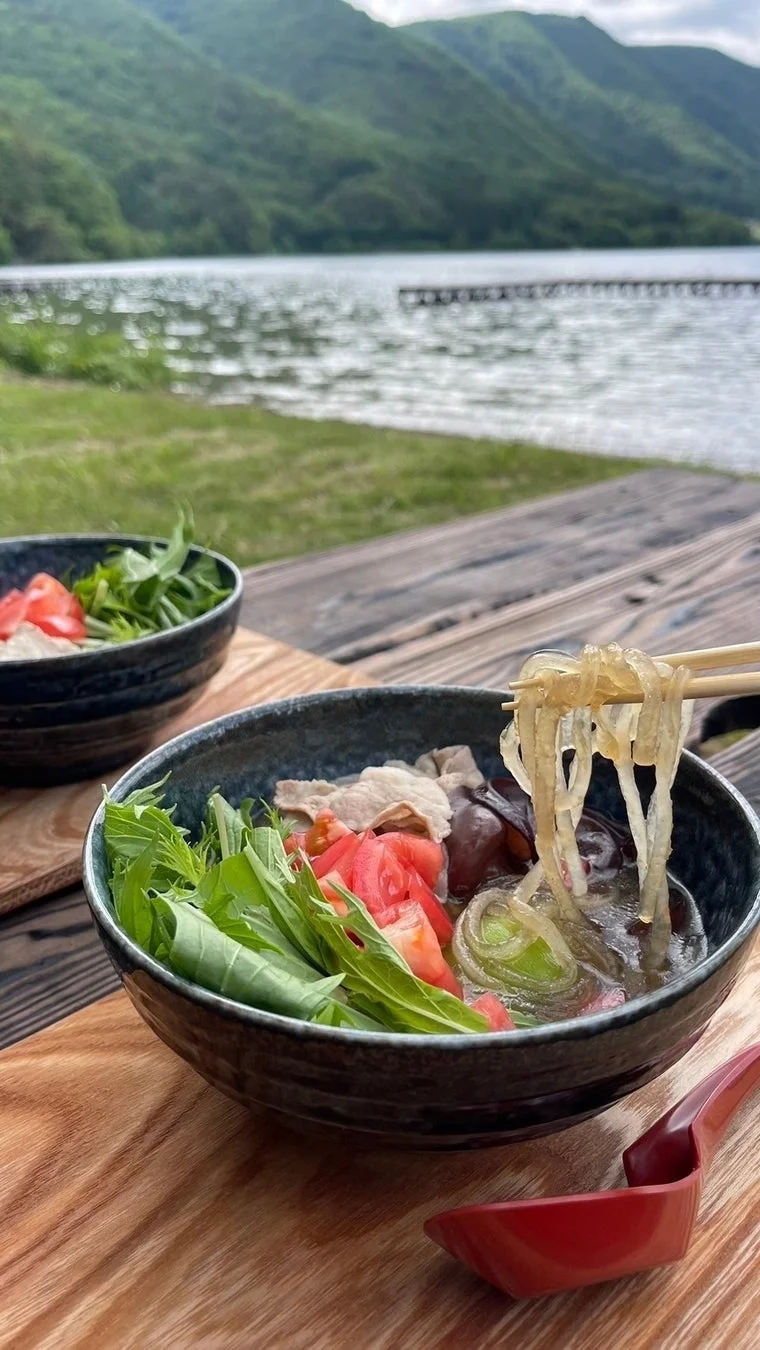 美しい湖畔を背景に、木製のテーブルでヘルシーな麺料理を楽しむアウトドアの様子