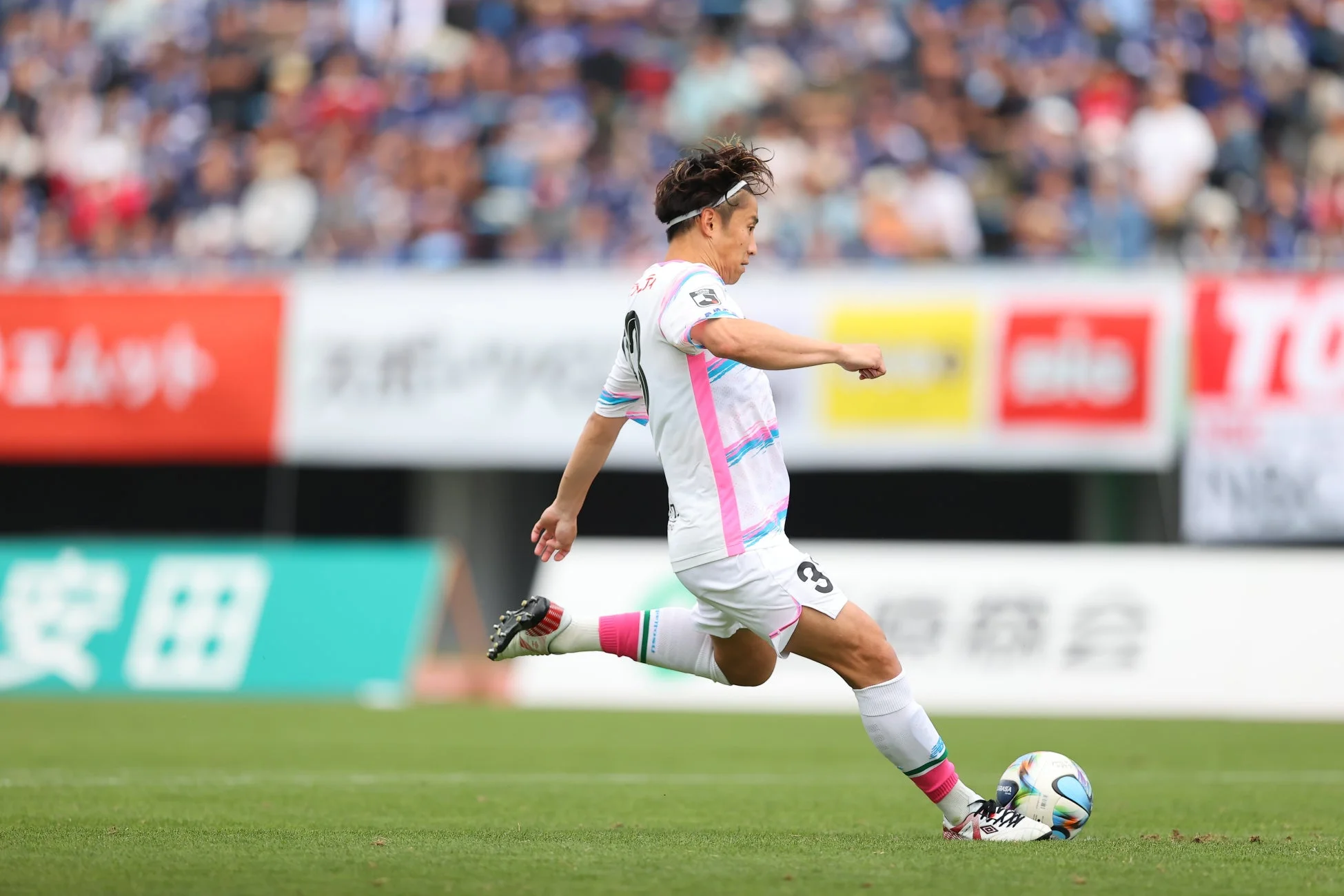 白いユニフォームを着たサッカー選手が、緑の芝生の上でボールを蹴る瞬間を捉えた写真です。背番号8の選手が力強くボールを蹴り込んでおり、背景には観客席とスポンサー看板が見えます。