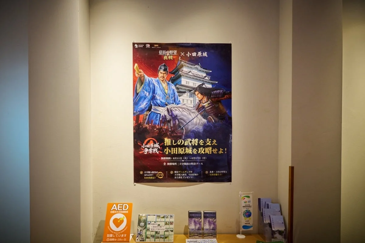 壁に掲示された「信長の野望」と「小田原城」のコラボイベントポスターがメイン。武将のイラストと小田原城が描かれ、イベント期間、割引、限定グッズ、甲冑撮影などの詳細が記されている。下部にはAED設置の案内も見える。