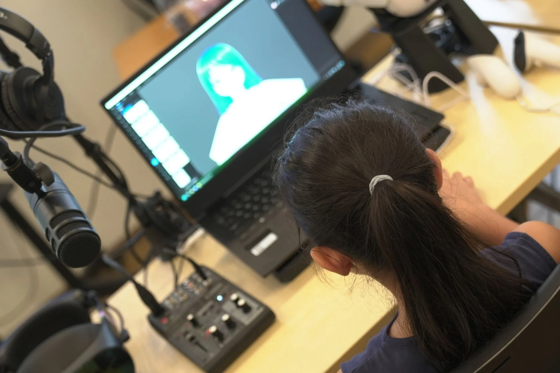デスクに向かい、デジタルアートや3Dモデルが映し出されたノートパソコンの画面を見つめる子供の後ろ姿。マイクやヘッドホン、オーディオミキサーが置かれ、コンテンツ制作や学習環境を示唆しています。