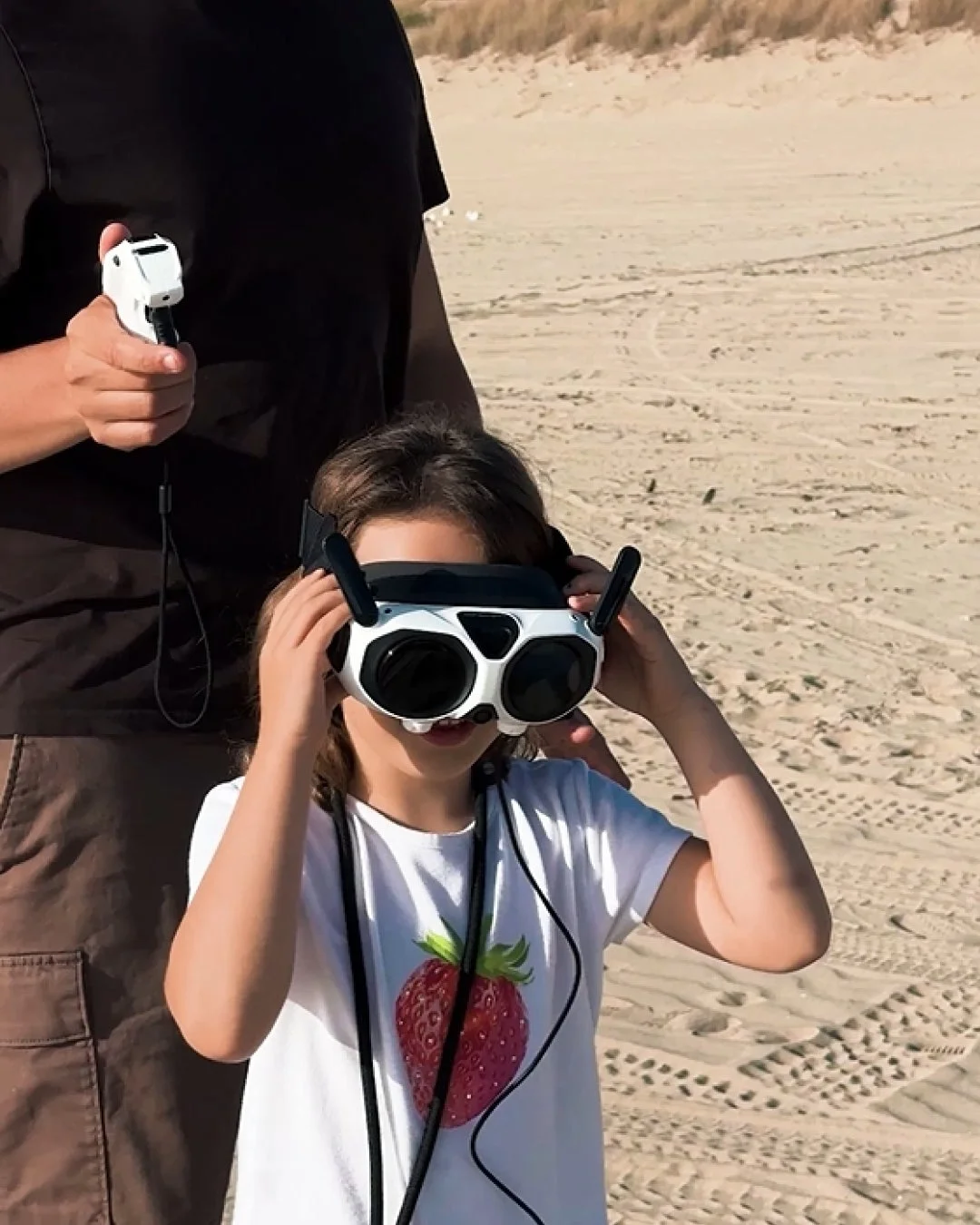 砂浜で、子供がFPVゴーグルを装着し、大人がドローンコントローラーを持っている様子。