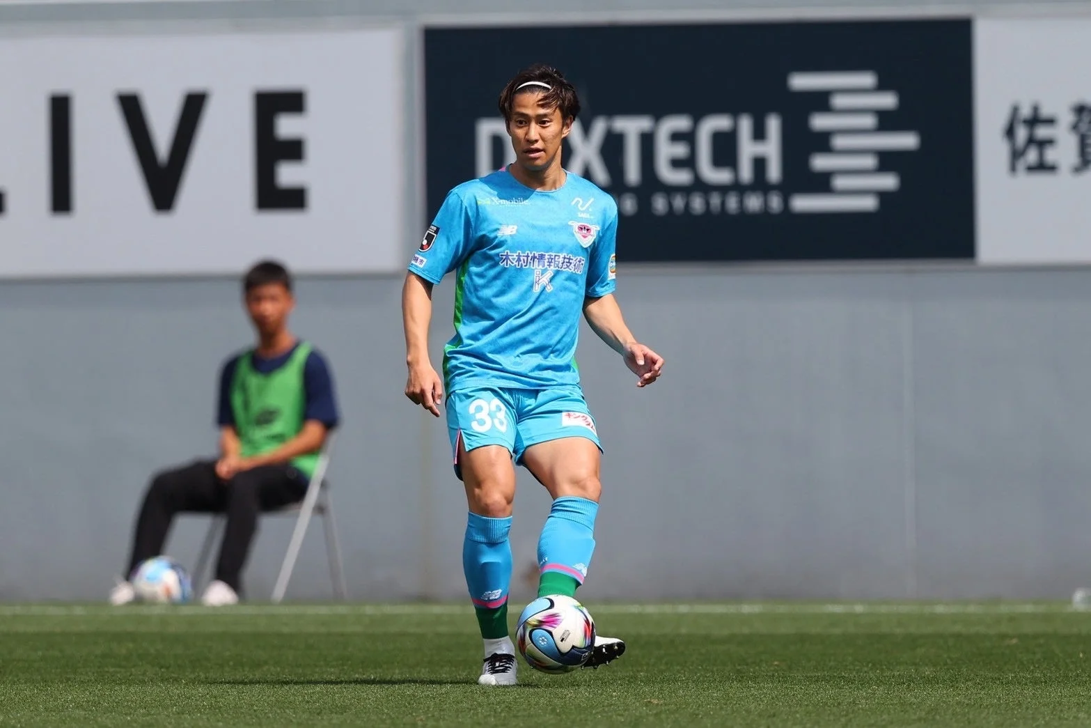 背番号33の選手が青いユニフォームを着て、緑の芝生の上でサッカーボールをドリブルしています。背景にはスポンサーの看板が見えます。