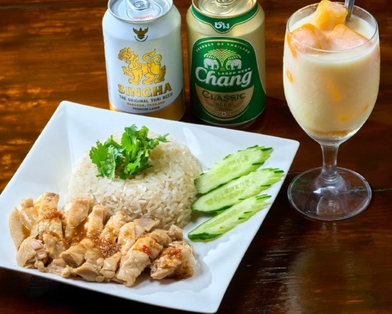 タイ料理のカオマンガイがメインの食卓。シンハービールとチャーンビールの缶、マンゴーのデザートドリンクが並ぶ