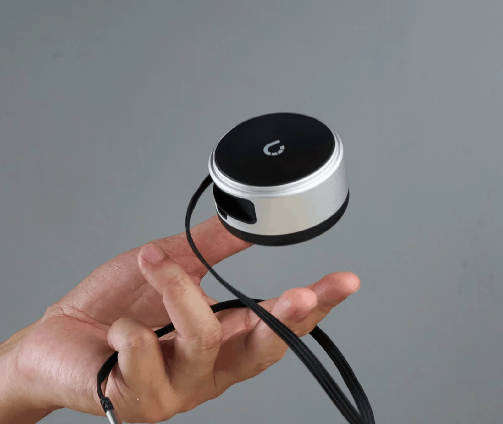 プレーンな灰色の背景をバックに、手が小型の円形電子デバイスを指で挟んで持っている様子。デバイスはシルバーとブラックで、上部にはU字型のロゴがあり、黒いコードが接続されています。