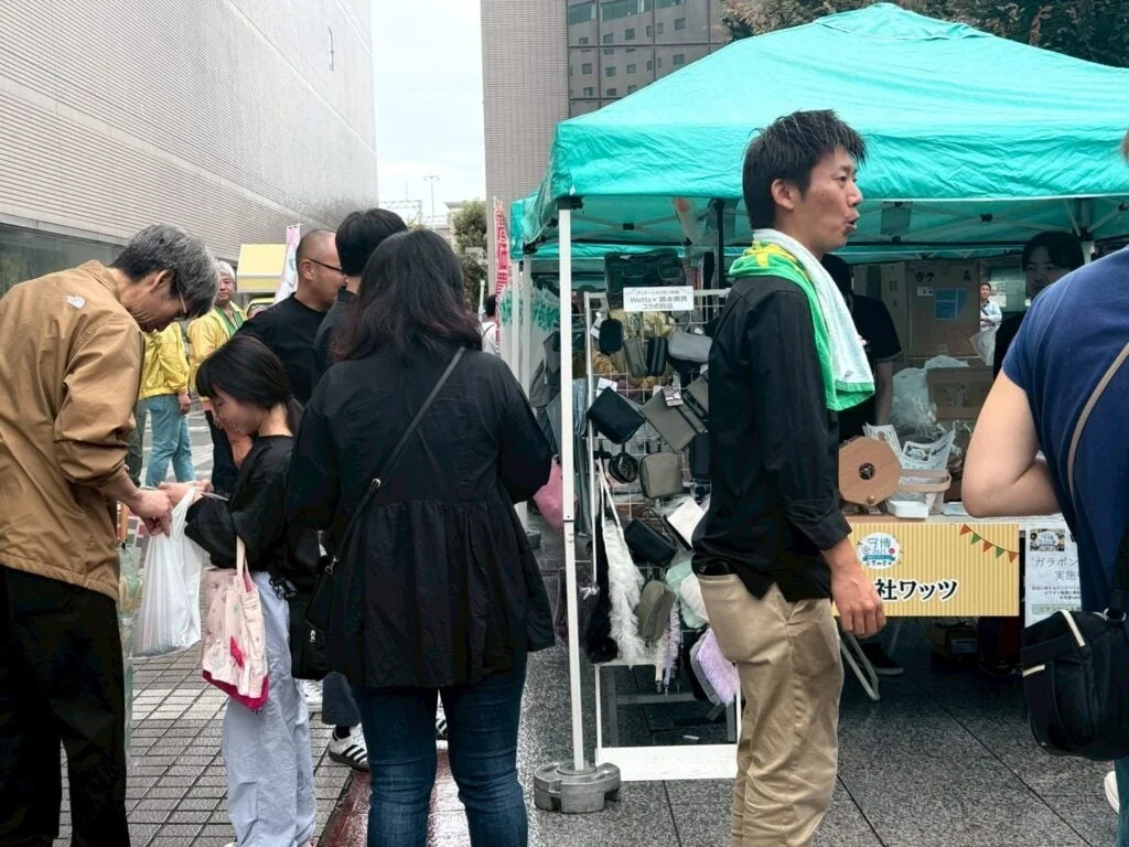屋外で開催されているイベントや市場の様子が写っています。複数の人々がブースの周りに集まっており、商品を見たり、会話したりしています。緑色のテントのブースでは「Watts × 岡本無線 コラボ企画」や「ガラポン実施」と書かれた看板が見られ、活気ある雰囲気が伝わってきます。
