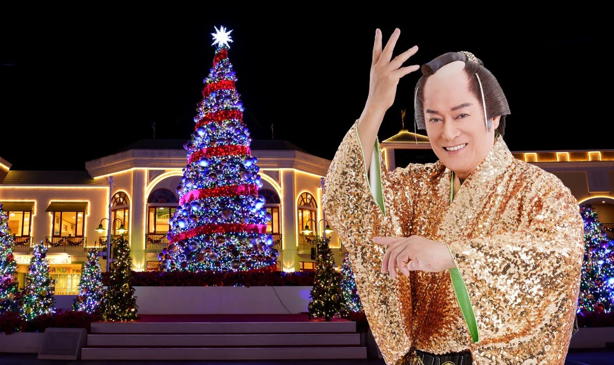 豪華なスパンコールの金色着物を着た日本人男性が、笑顔でポーズをとっています。背景には、鮮やかにライトアップされた巨大なクリスマスツリーと建物があり、華やかで祝祭的な夜の雰囲気を醸し出しています。