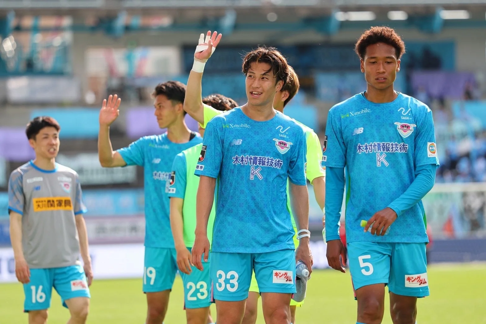 晴れた日のスタジアムで、水色とグレーのユニフォームを着た数人のサッカー選手が立っている様子。選手の一人が手を振っており、試合中か試合後の一コマと思われる。