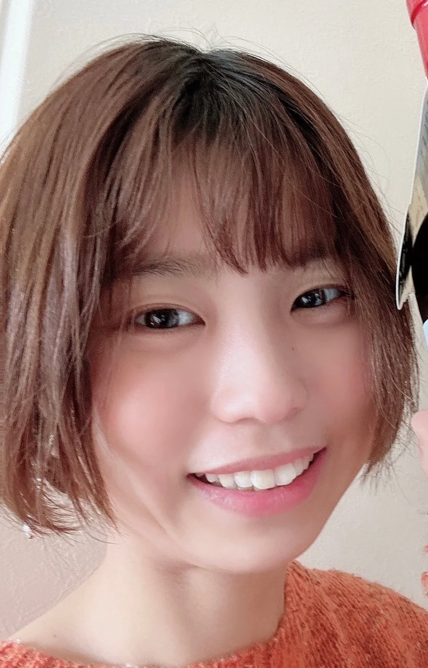 笑顔でカメラを見つめる若い女性のクローズアップポートレート。茶色のショートヘアで、明るいオレンジ色のニットを着ています。親しみやすく、楽しそうな表情をしています。