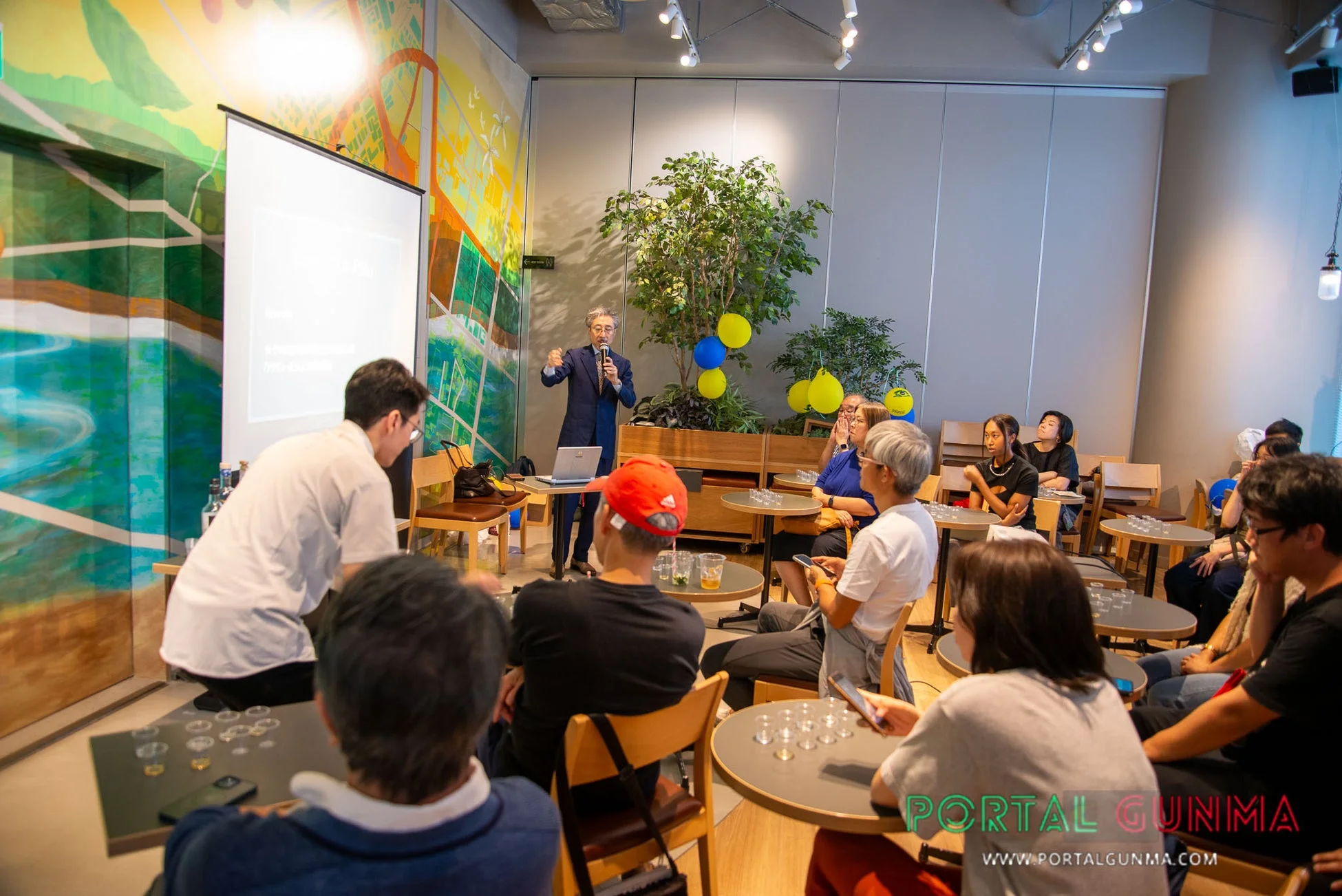 講演者がマイクを持ってプレゼンテーションを行い、集まった人々が熱心に耳を傾けている屋内のイベント風景