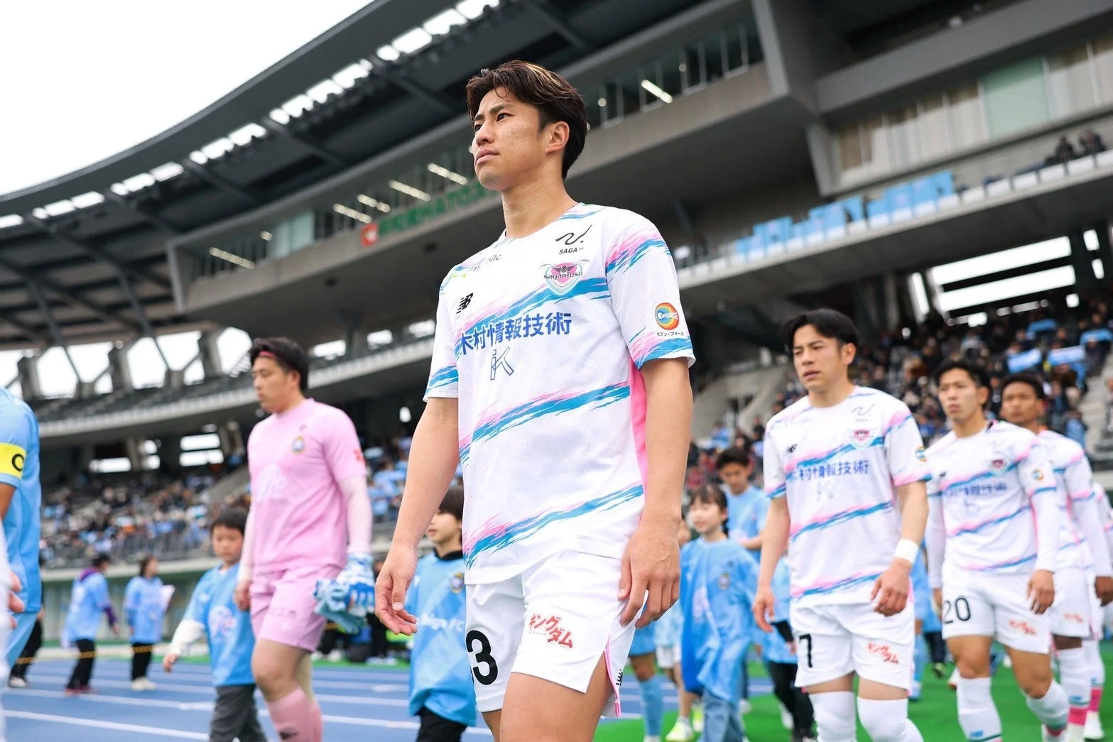 サッカーの試合前に選手たちがスタジアムのピッチに登場している様子を捉えた一枚です。白地にピンクと水色のストライプが入ったユニフォームを着た選手たちが、真剣な表情で歩いています。手前には背番号3の選手がおり、奥には他の選手や子供たちが続いています。