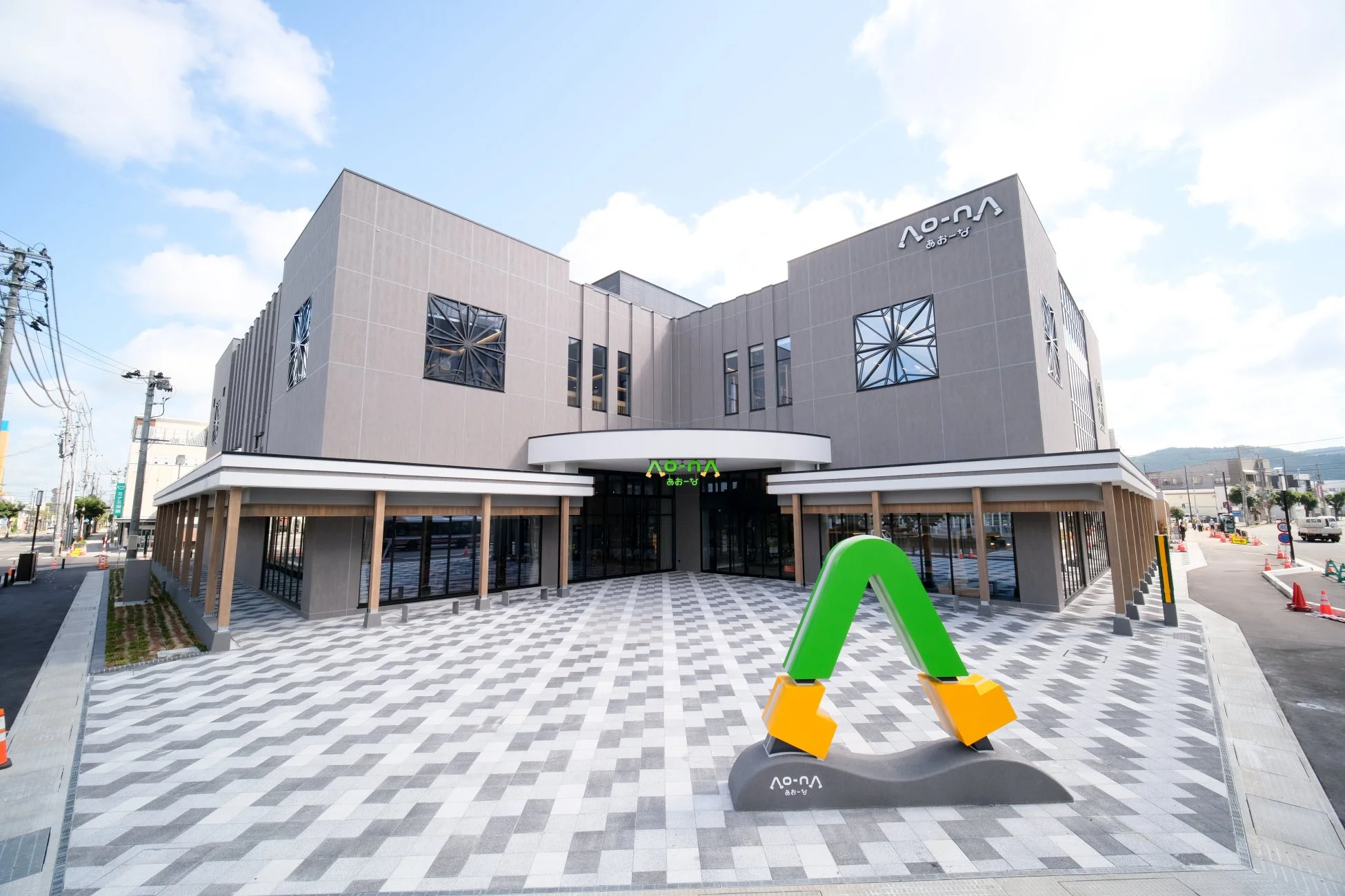 晴れた日に撮影された、モダンなデザインの「a-o-na あおーな」という名前の建物です。特徴的な緑色の「A」型モニュメントが広場に設置されており、施設の入り口にも同じロゴが見えます。
