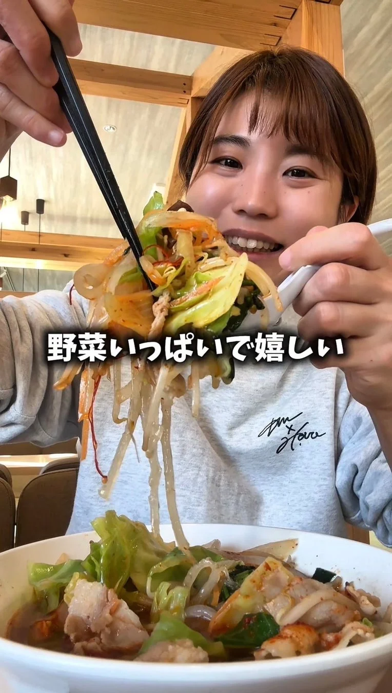 笑顔の女性が、箸とレンゲを使って野菜が山盛りの美味しそうな麺料理を食べている。画像には「野菜いっぱいで嬉しい」という文字があり、食事の満足感が伝わる。