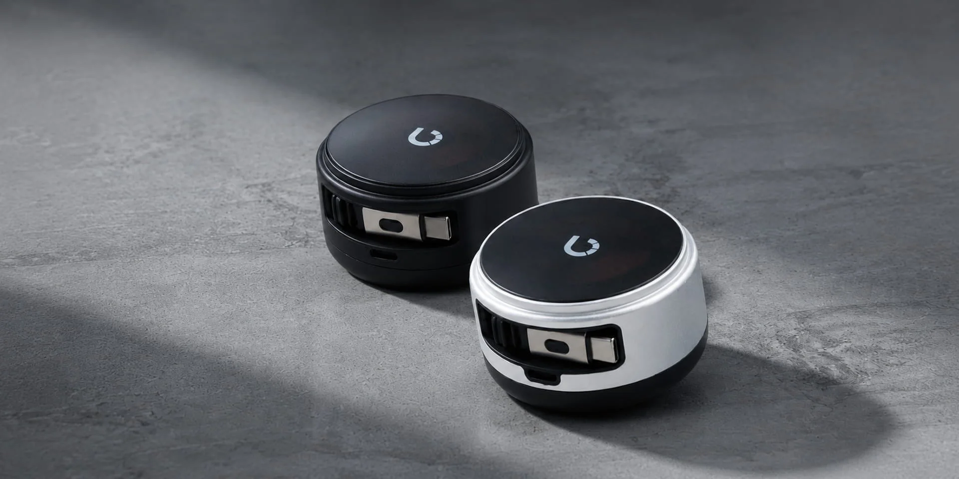 黒と白の2つの丸いコンパクトな電子機器が、灰色で質感のある表面に置かれています。それぞれの機器の上面には特徴的なロゴがあり、側面には金属製のクリップのような部分が見えます。
