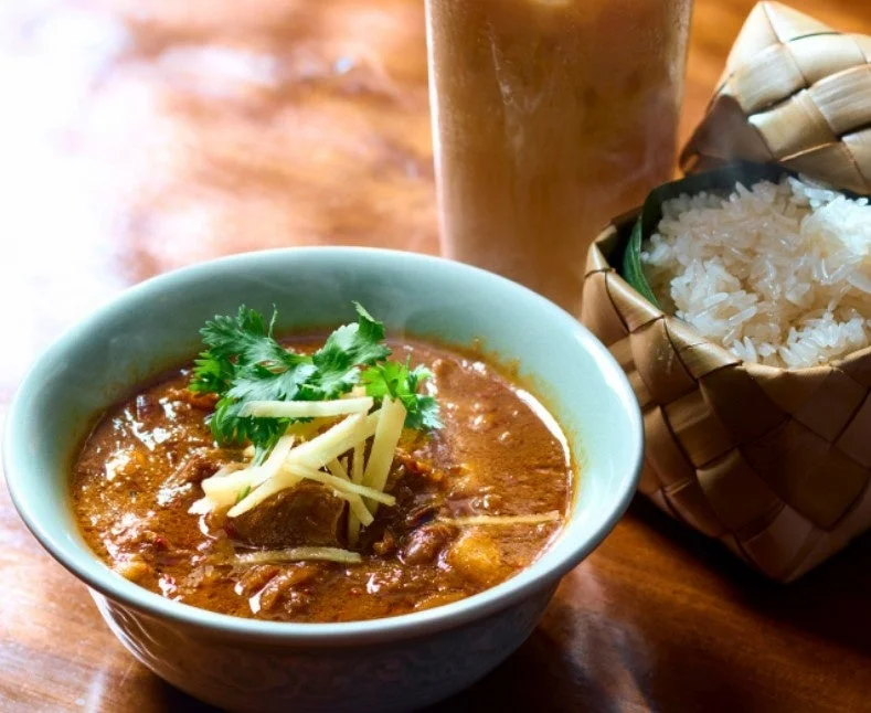 スパイシーなカレーと伝統的な竹かごに入ったもち米、飲み物が木製のテーブルに並べられたアジアンミール