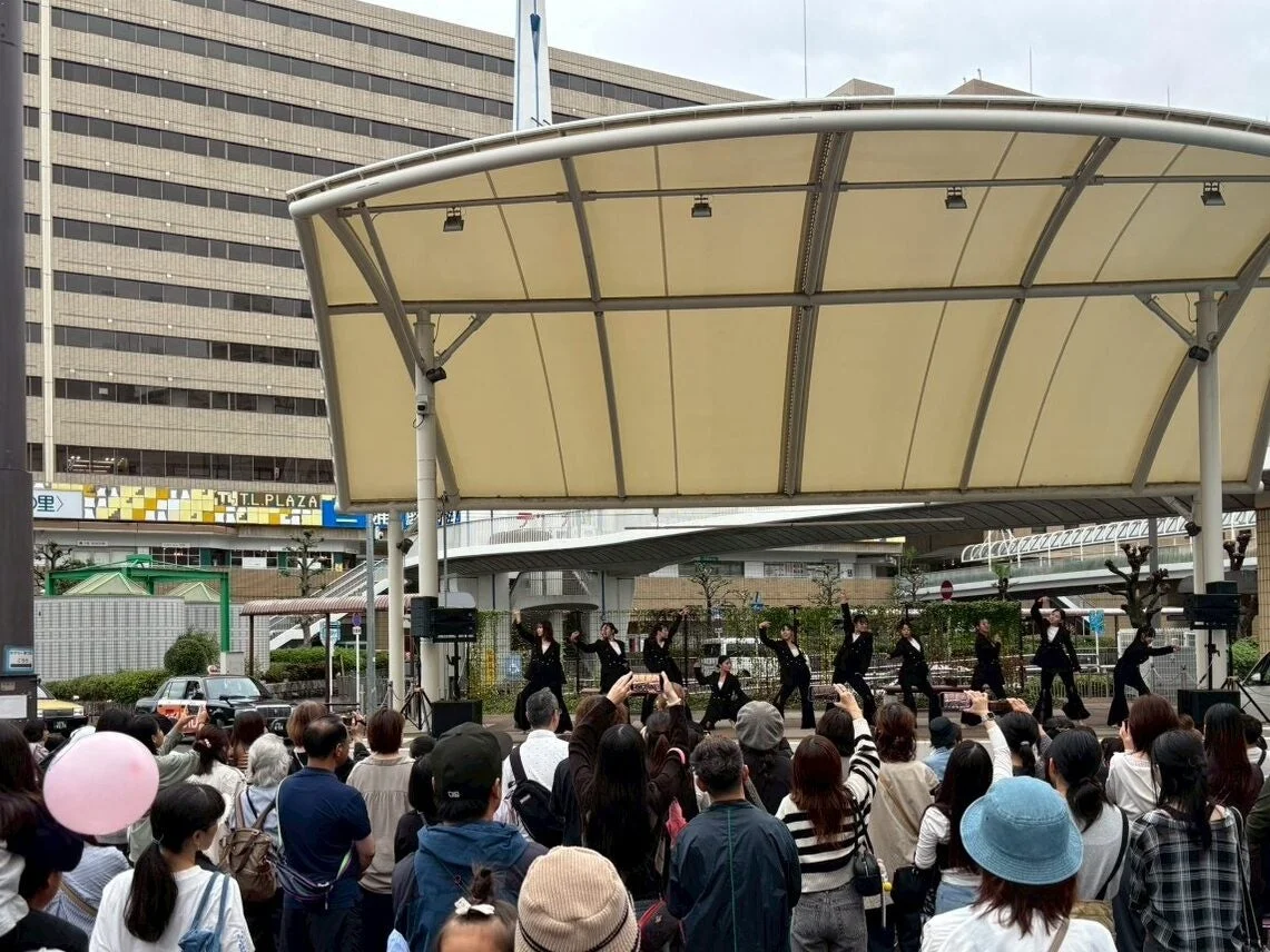 屋外のステージで、複数のダンサーが観客の前でパフォーマンスを披露しています。多くの人々がその様子を鑑賞しており、中にはスマートフォンで撮影している人もいます。