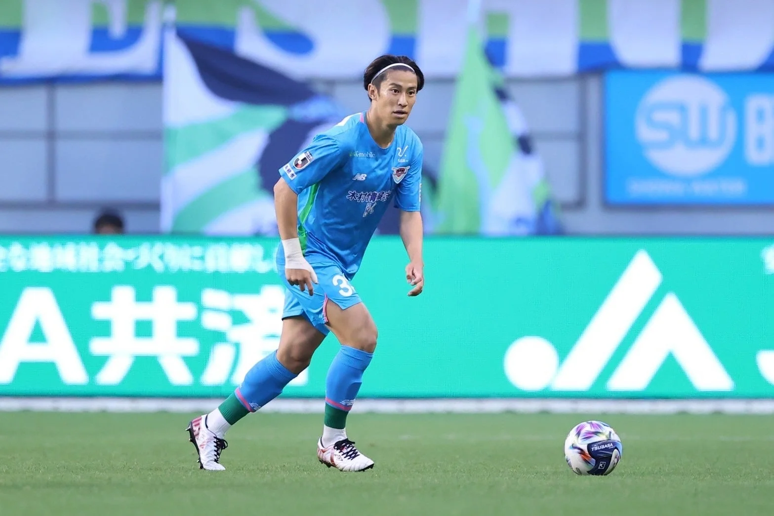 青いユニフォームを着たサッカー選手が、緑の芝生の上でボールの近くに立っています。スタジアムのLED広告ボードを背景に、試合中の真剣な表情が印象的です。