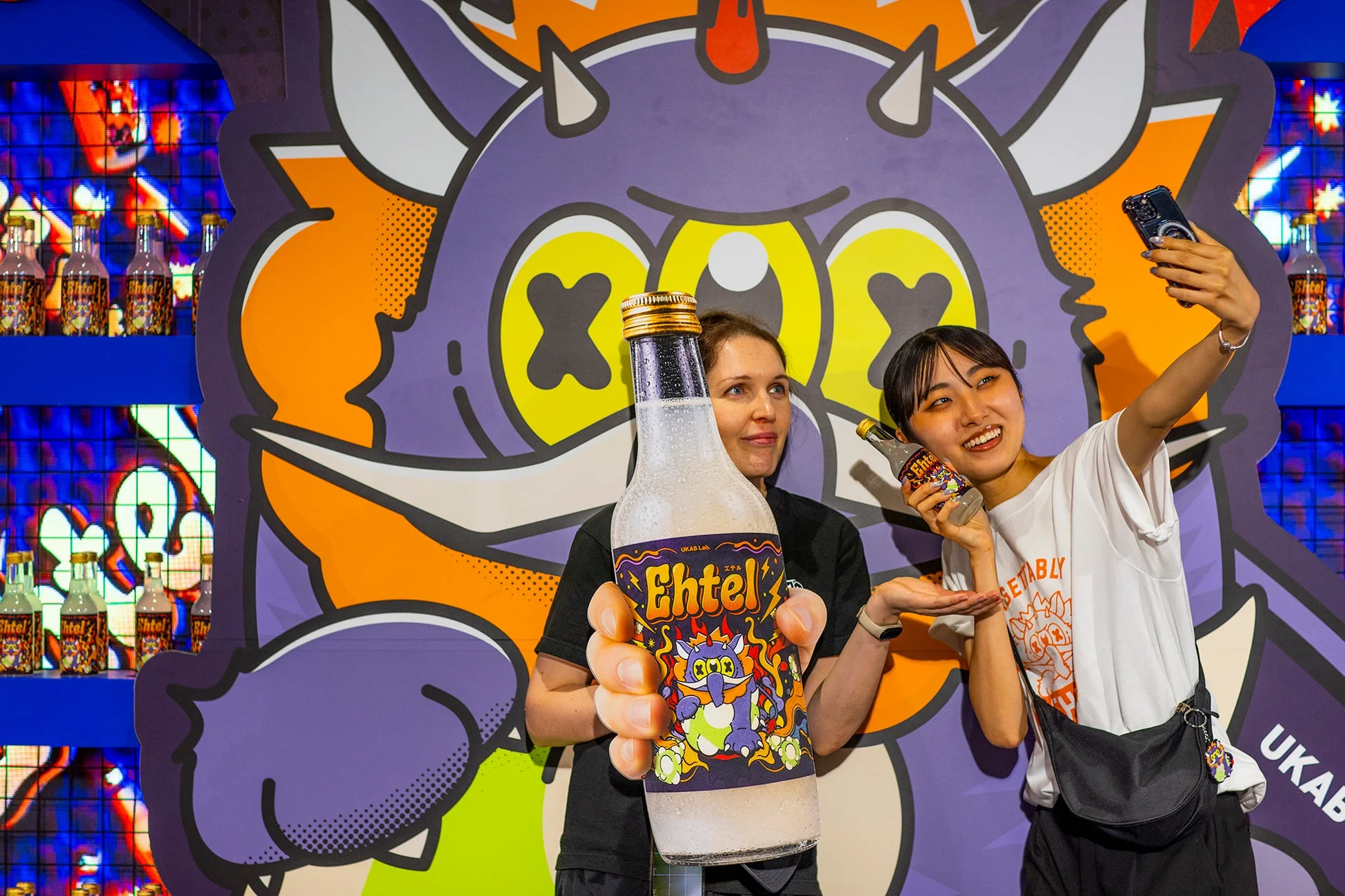 カラフルなモンスターのイラストが描かれた背景の前で、2人の女性が「Ehtel」の飲料ボトルを持って楽しんでいる