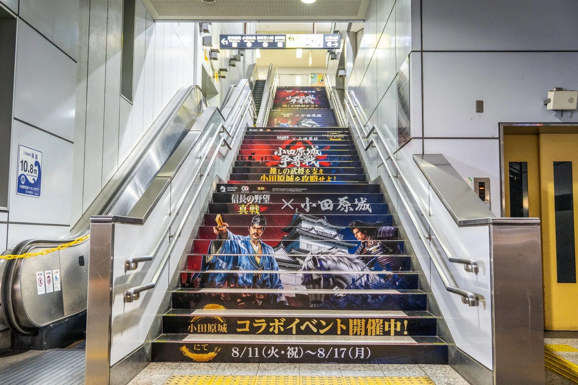 小田原駅構内の階段に、ゲーム「信長の野望・真戦」と「小田原城」のコラボイベント広告が大きく掲示されています。武将や城のイラストが階段の蹴上部分に描かれ、イベント期間や内容が告知されており、目を引くデザインです。