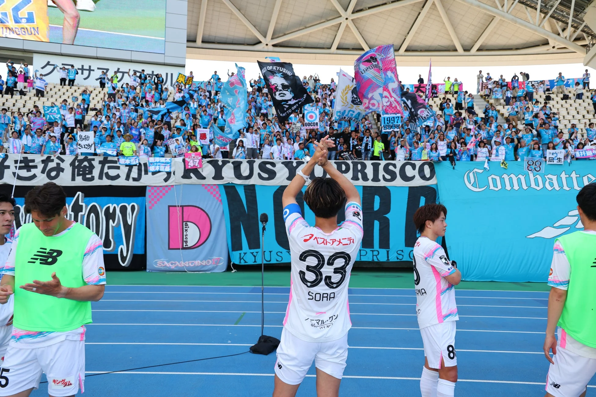 サッカーの試合会場で、選手がサポーターの声援に応えている様子。多くの観客が青いユニフォームや旗を掲げて応援し、スタジアム全体に熱気と一体感が満ちている。背番号33の選手が拍手で感謝を示している。